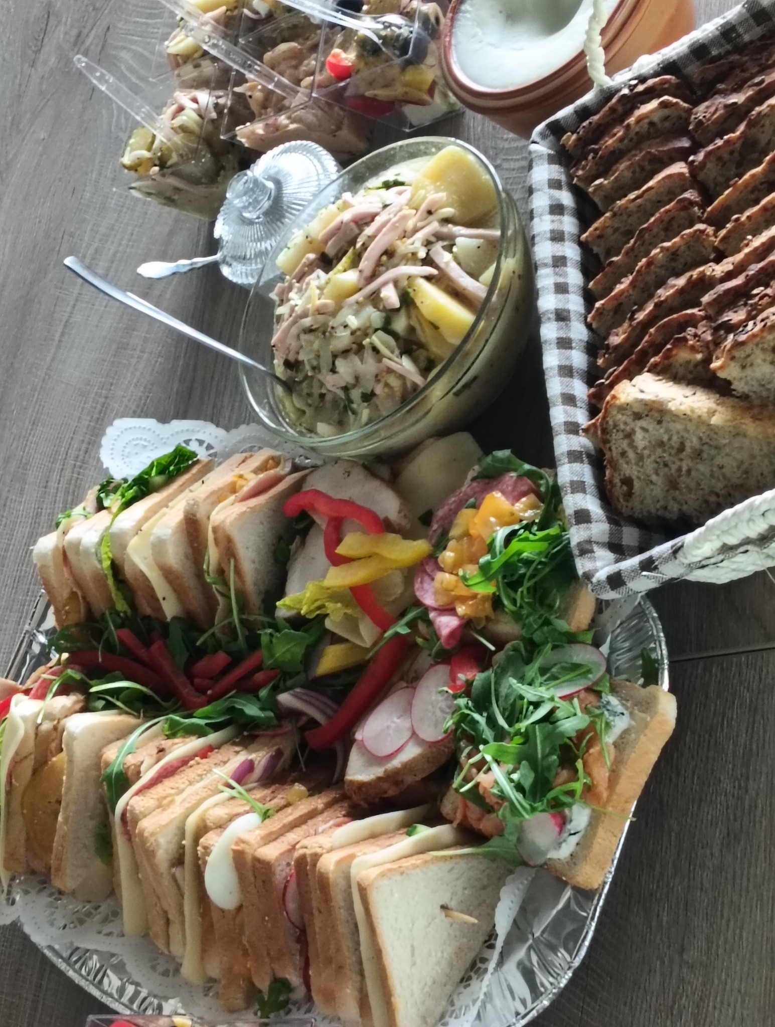 Bogato zastawiony stół: kanapki na srebrnej tacy, sałatki w szklanych misach i pieczywo w koszyku z materiału w kratę. Idealne na imprezę.