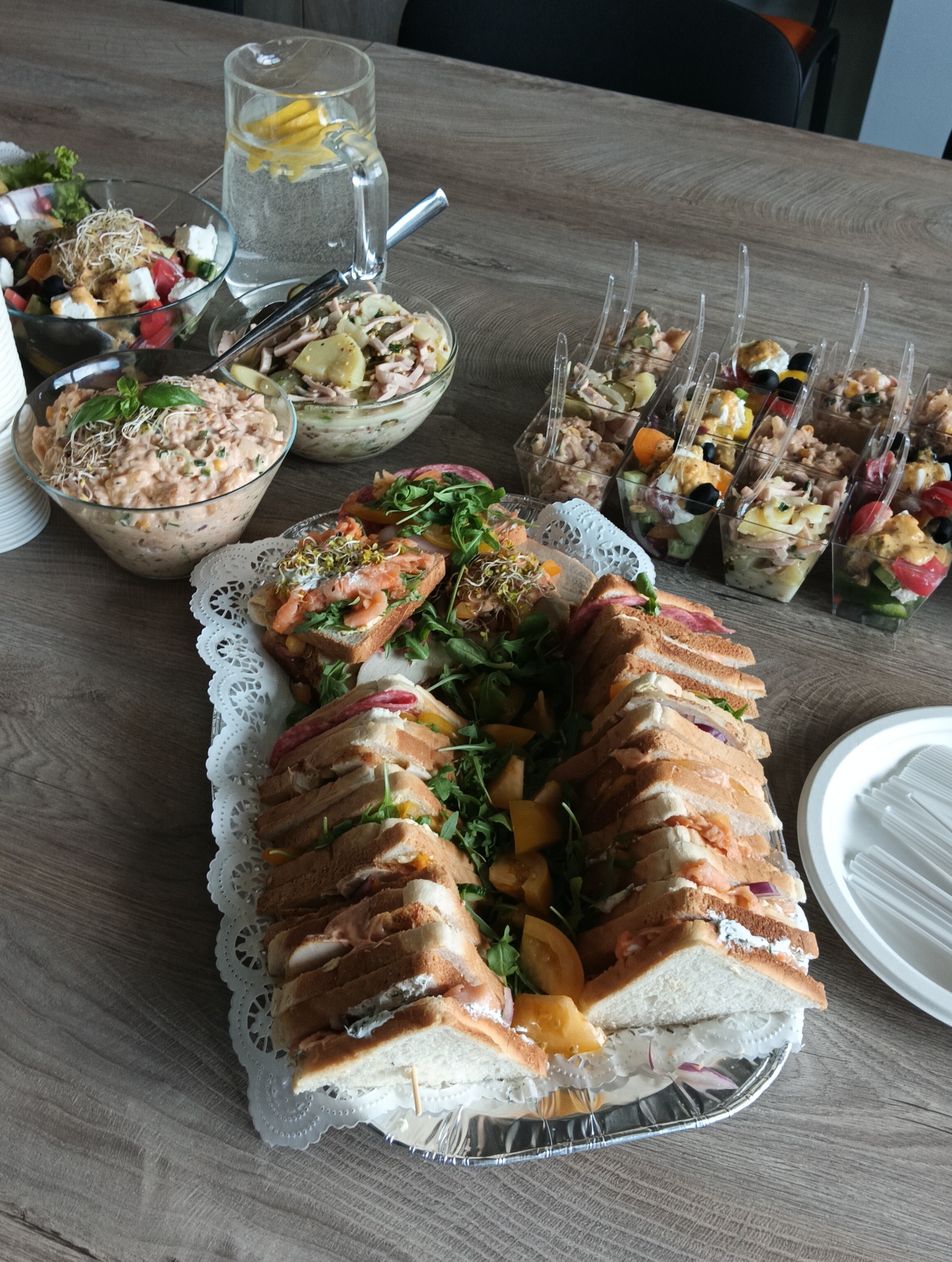 Stół z cateringiem: kanapki na ozdobnej tacy, sałatki w miskach i plastikowych kubeczkach, dzbanek z wodą i cytryną. Widok z góry, oświetlenie naturalne.