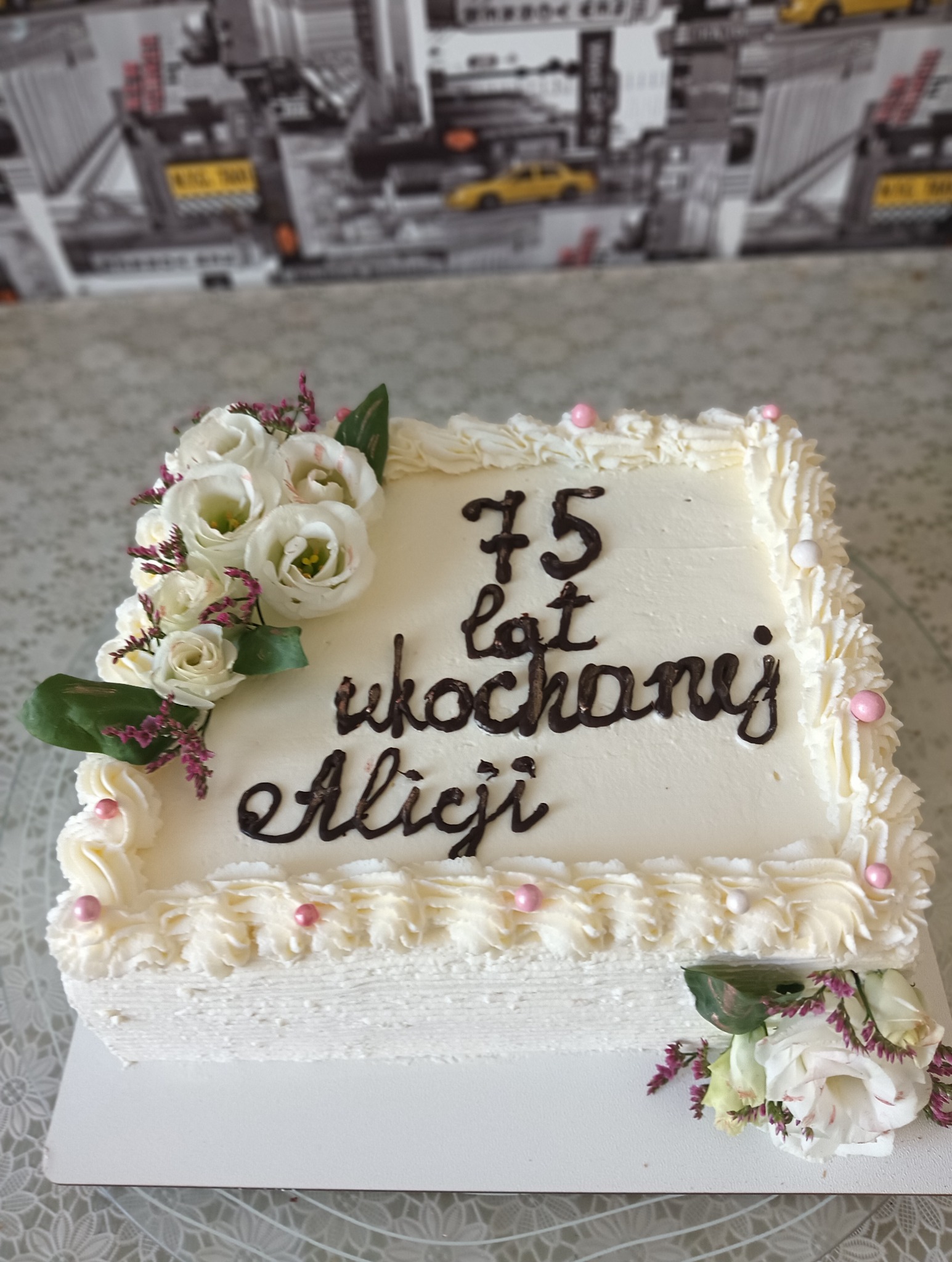 Kwadratowy tort z kremem i kwiatami w Olsztynie. Napis z czekolady '75 lat ukochanej Alicji'. Dekoracja z białych róż i różowych koralików.