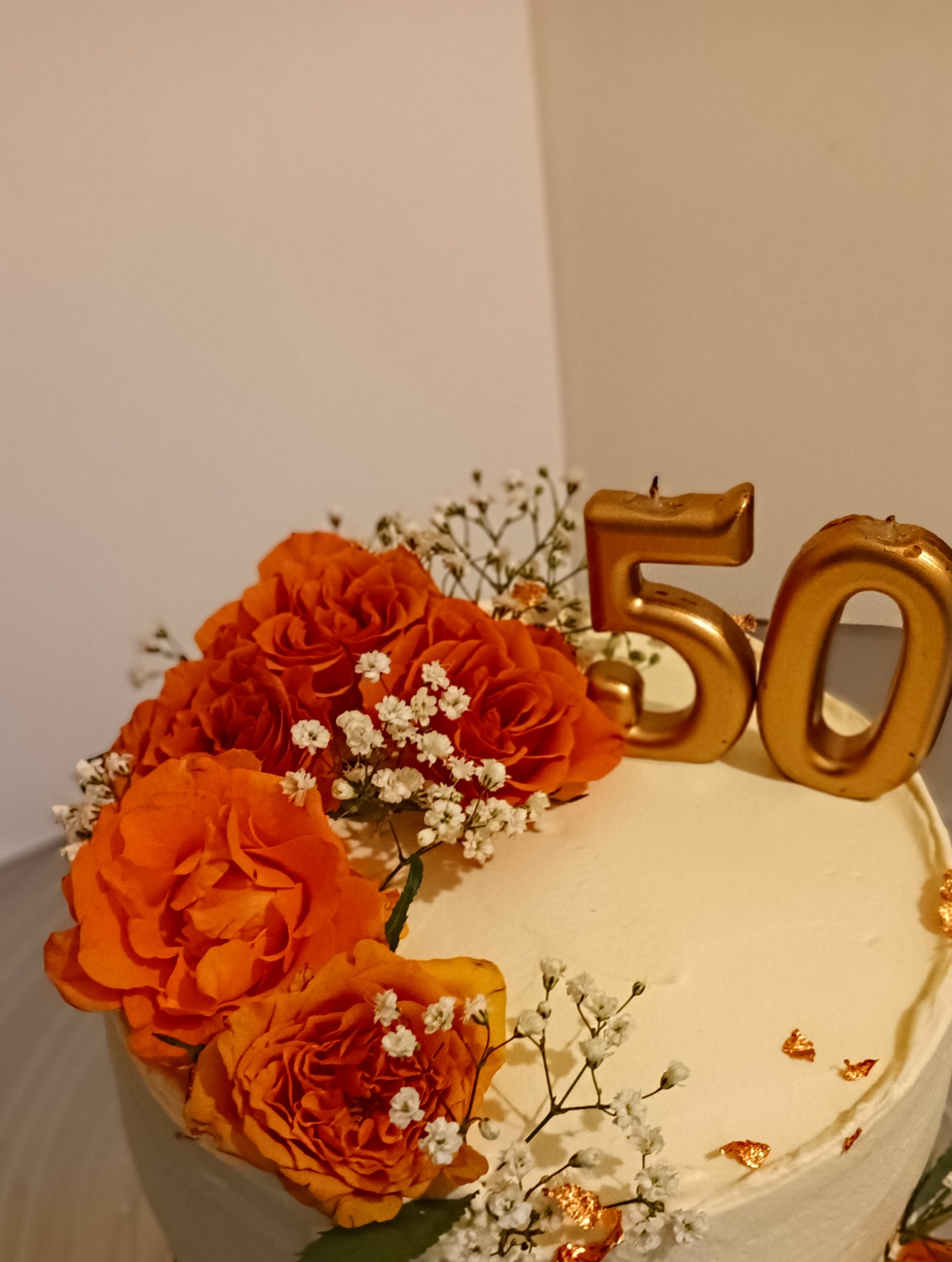 Tort urodzinowy z kremem, pomarańczowymi różami i świeczkami w kształcie cyfr '50', dekorowany złotymi płatkami.