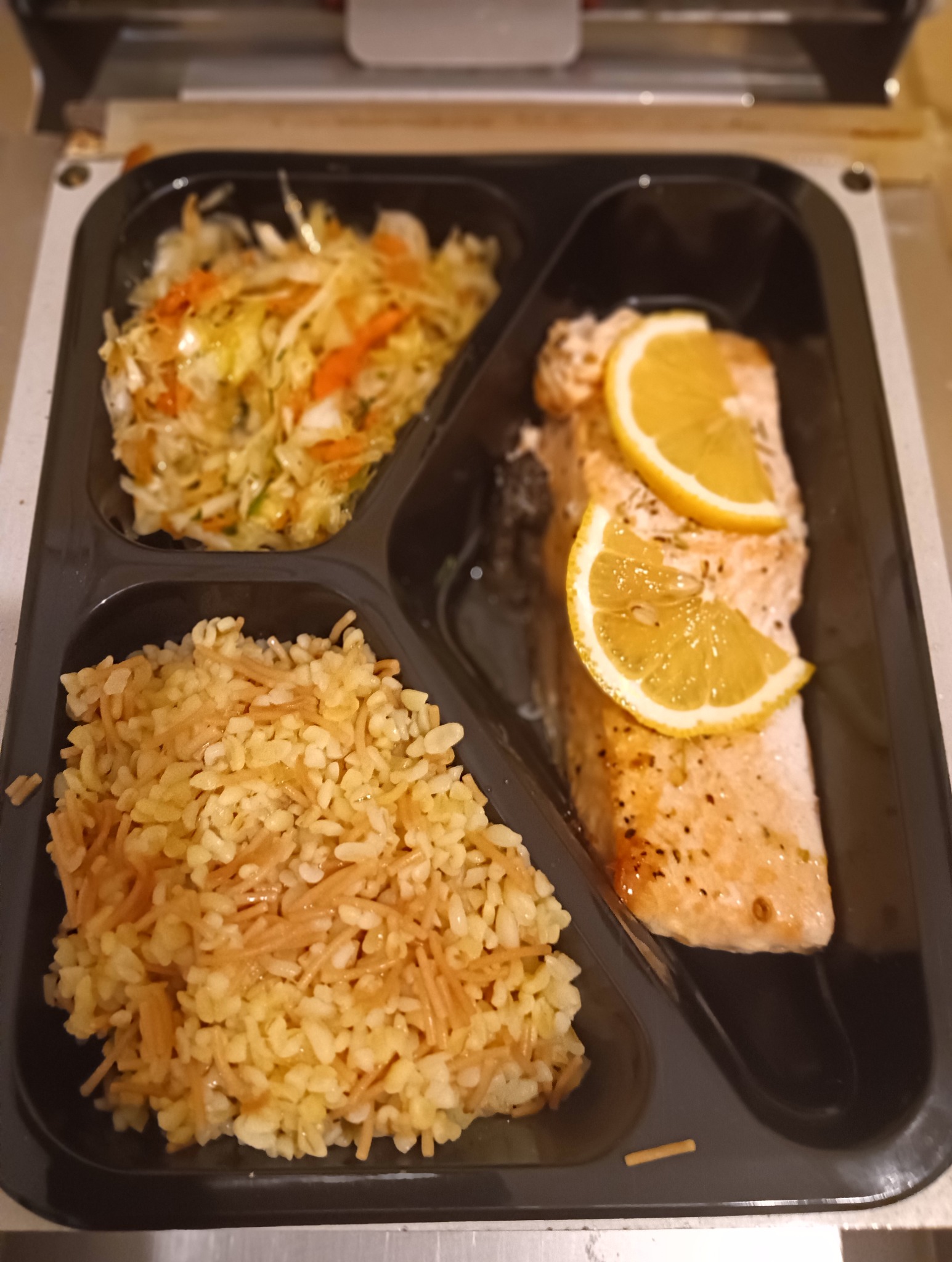 Zestaw obiadowy w czarnym pojemniku: ryba z cytryną, ryż z makaronem i surówka. Gotowy posiłek cateringowy, widok z góry.