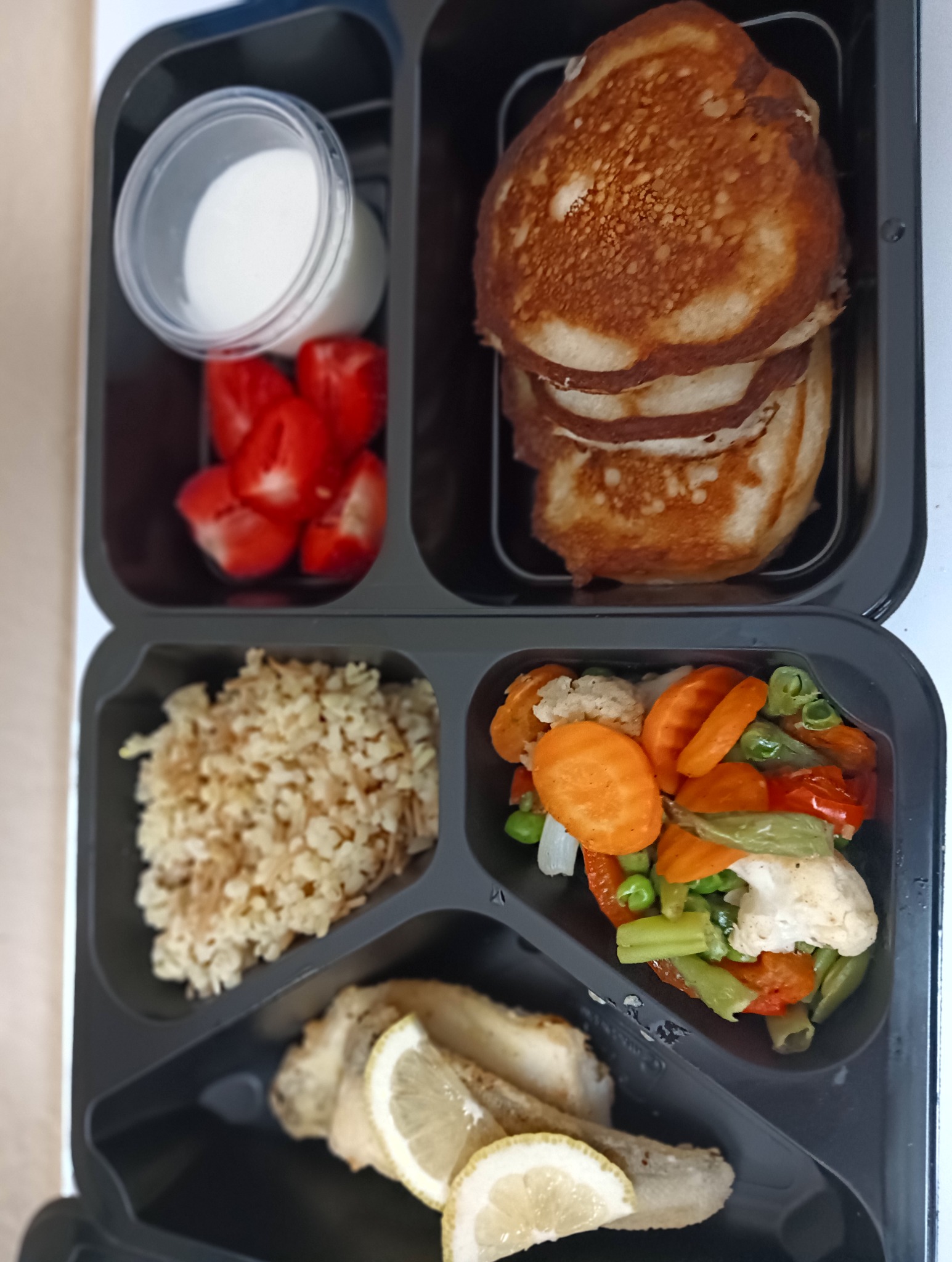 Czarny pojemnik cateringowy z pancakes, sosem, truskawkami, kaszą i warzywami. Ryba z cytryną w dolnej przegródce. Zdrowy, zbilansowany posiłek.