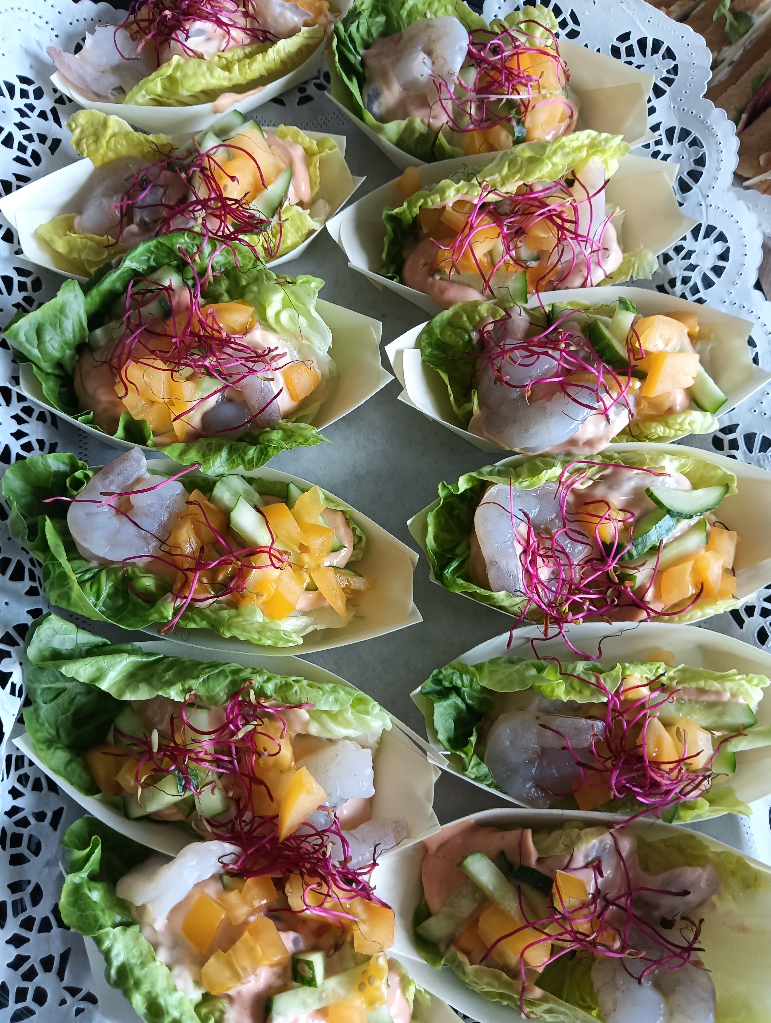 Egzotyczne przystawki cateringowe: krewetki, mango, ogórek i sałata w łódeczkach z jasnego drewna, udekorowane kiełkami buraka. Elegancki i świeży finger food.