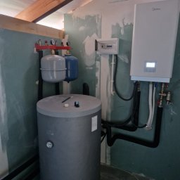 Aqua tech - Szary zbiornik buforowy z osprzętem, obok biała pompa ciepła Midea na ścianie. Widoczne rury, zawory i skrzynka elektryczna na tle surowych ścian.