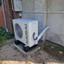 Zamontowana pompa ciepła Kaisai na betonowej podstawie przy ścianie budynku z widocznymi rurami instalacyjnymi i skrzynką elektryczną na elewacji.