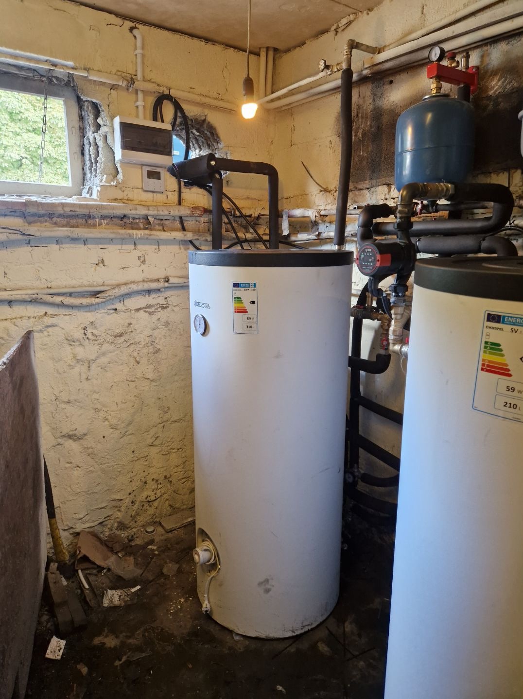 Bojlery Kospel z czarnymi rurami w piwnicy z oknem. Widoczne etykiety energetyczne, instalacja wodna, pompa obiegowa i wisząca żarówka. Ściany z surowej cegły.