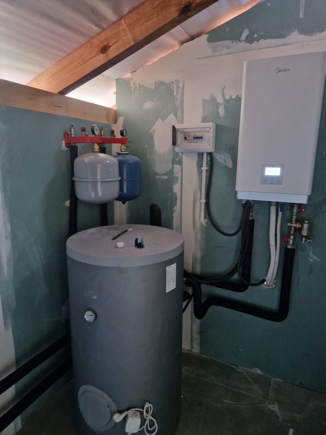 Szary zbiornik buforowy z osprzętem, obok biała pompa ciepła Midea na ścianie. Widoczne rury, zawory i skrzynka elektryczna na tle surowych ścian.