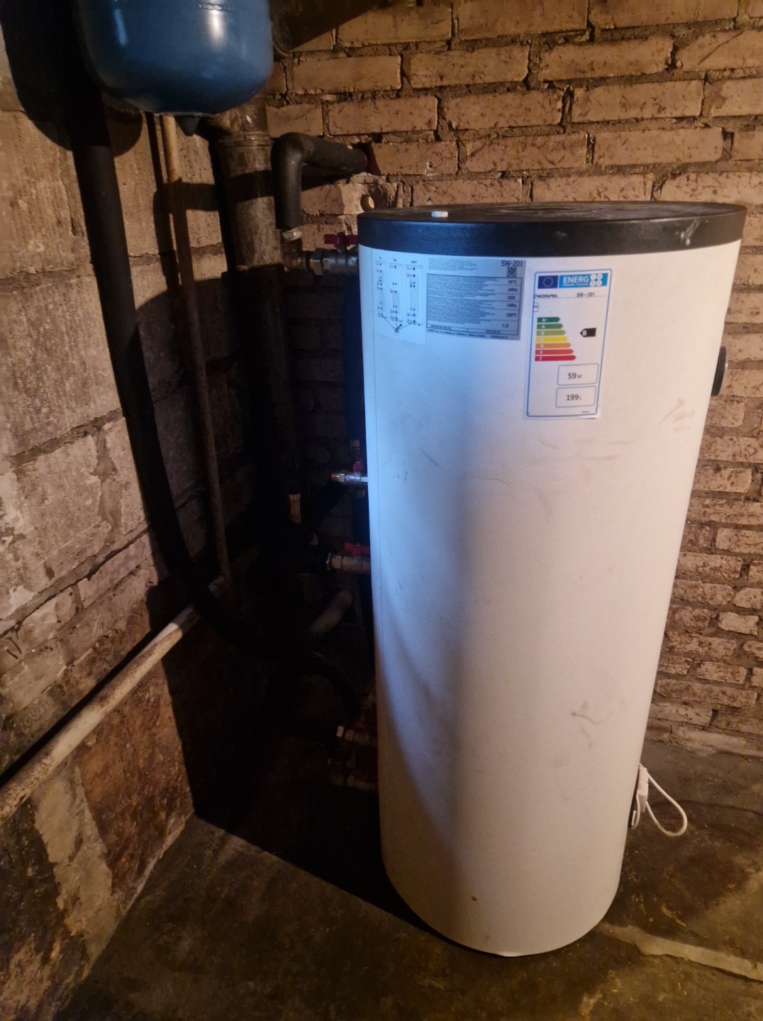 Bojler z etykietą energetyczną w kotłowni z ceglaną ścianą. Widoczne rury i instalacja. Oświetlenie z góry podkreśla fakturę cegieł.