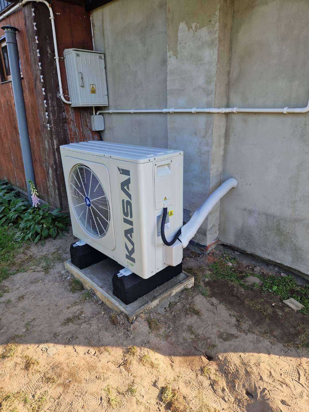 Zamontowana pompa ciepła Kaisai na betonowej podstawie przy ścianie budynku z widocznymi rurami instalacyjnymi i skrzynką elektryczną na elewacji.