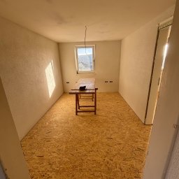 OMI Kacper Omylak - Pokój z podłogą wyłożoną płytami OSB, drewniany stół pośrodku, okno z widokiem na zewnątrz. Przygotowanie do dalszych prac wykończeniowych.