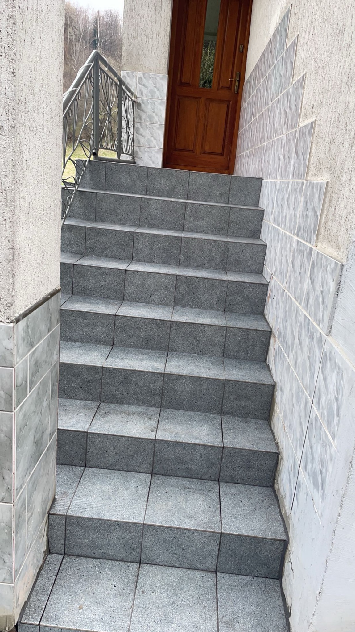 Zewnętrzne schody wyłożone szarymi płytkami prowadzą do drewnianych drzwi wejściowych. Fuga w kolorze szarym. Ściana obłożona białymi, kwadratowymi płytkami.