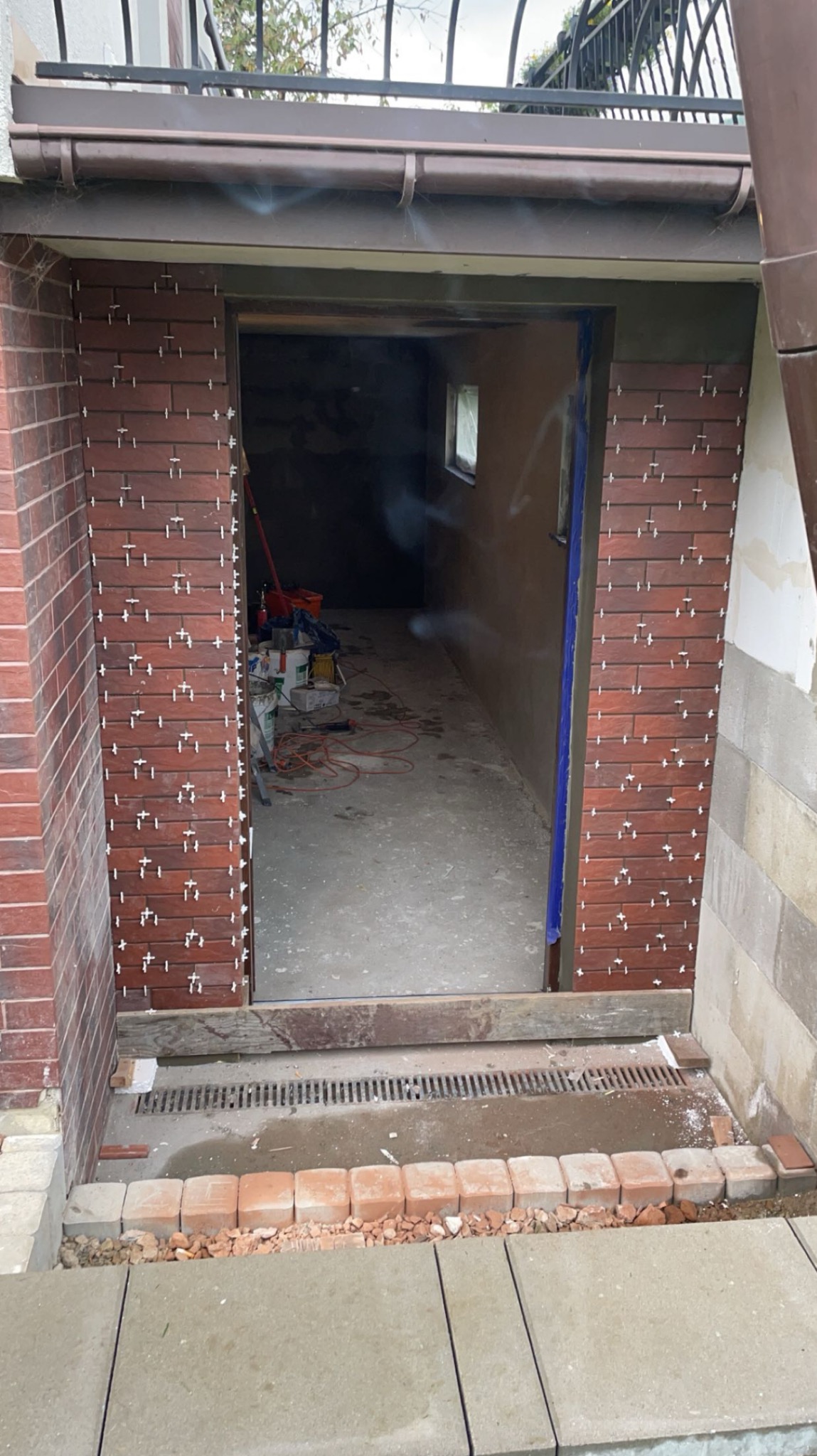 Kładzenie cegieł klinkierowych wokół wejścia do budynku. Widoczne krzyżyki dystansowe, narzędzia w tle i elementy wykończeniowe. Prace glazurnicze w toku.