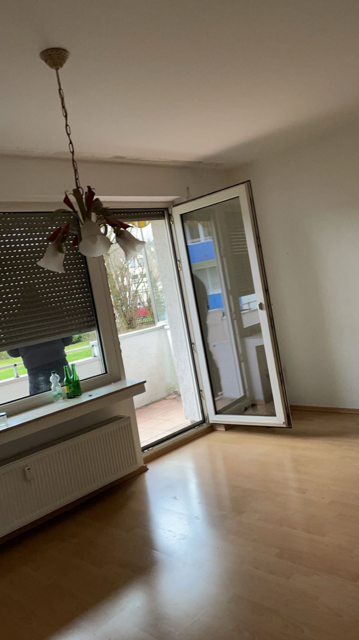 Jasny pokój z parkietem, otwartym balkonem i oknem z roletą. W tle widoczny balkon sąsiada. Na parapecie stoją dwie butelki. Lampa zwisa z sufitu.