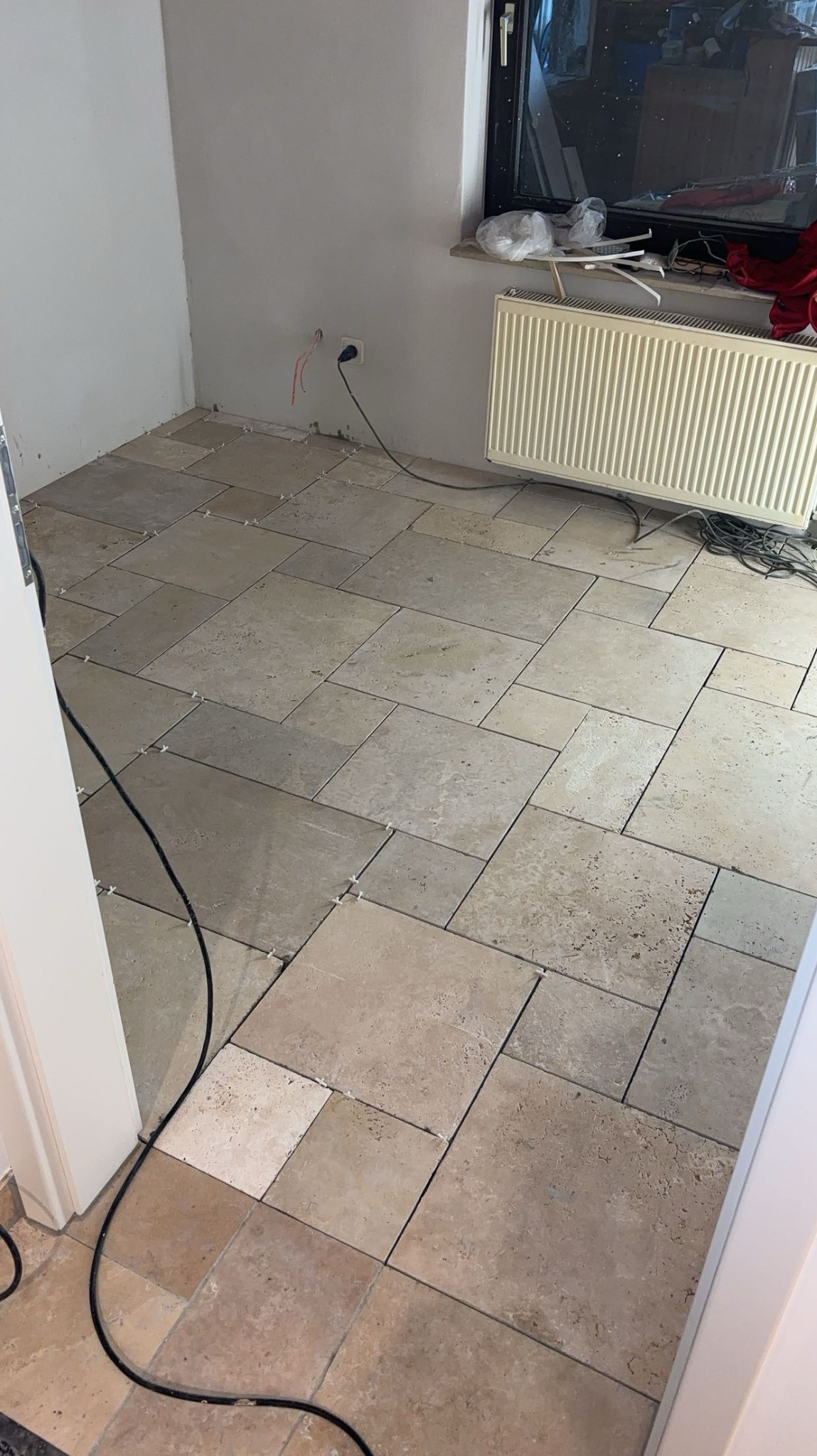 Układanie płytek podłogowych w pomieszczeniu, widoczne krzyżyki dystansowe, kabel elektryczny przy ścianie, grzejnik pod oknem, beżowe płytki w różnych odcieniach.