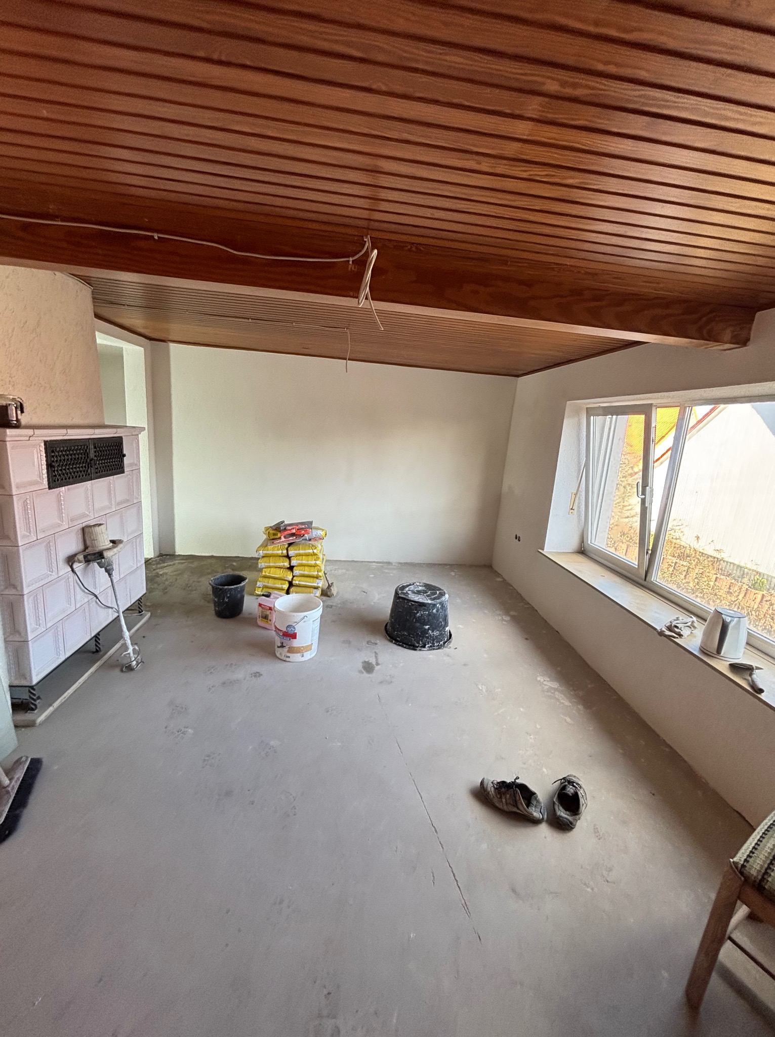 Wnętrze w trakcie remontu: worki z zaprawą, wiaderka, piec kaflowy, drewniany sufit. Widoczne ślady prac remontowych na podłodze. Dziennie światło wpada przez okno.
