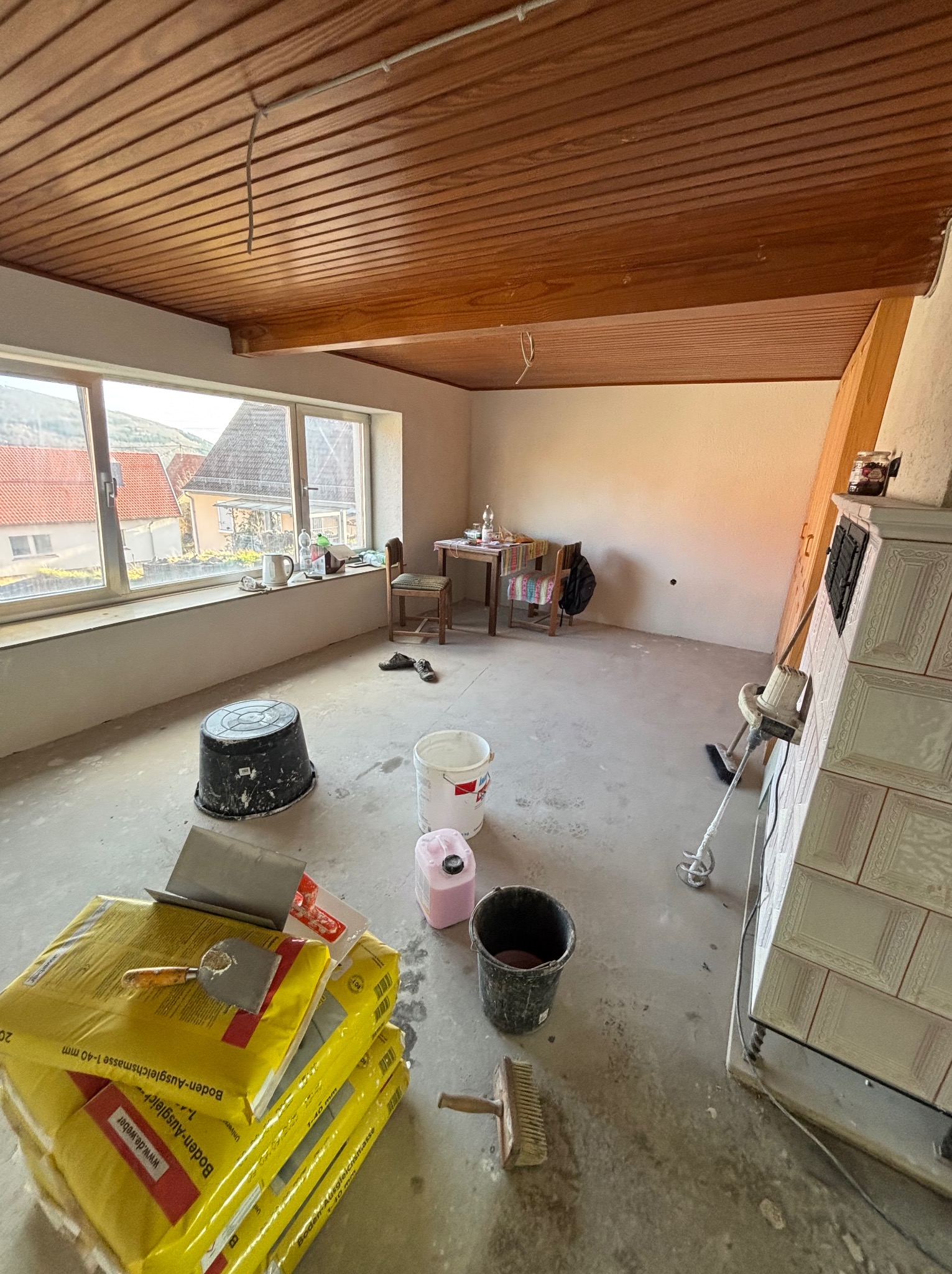 Przygotowania do wylewki samopoziomującej: worki z masą, wiadro, mieszadło i narzędzia w surowym wnętrzu z drewnianym stropem i oknem na wiejski krajobraz.