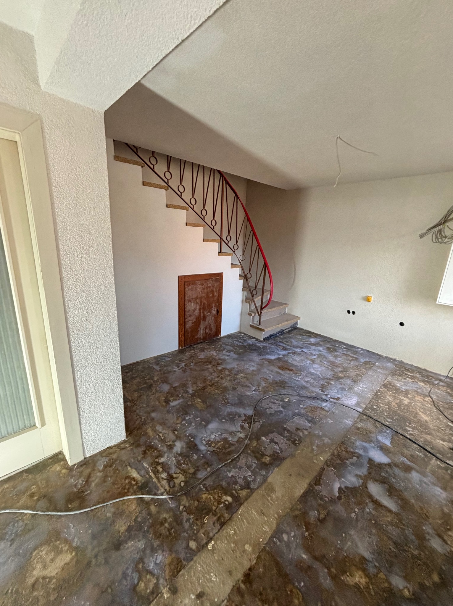 Wnętrze w trakcie remontu: schody z czerwoną balustradą, podłoga z resztkami kleju, ściany przygotowane do malowania, widoczne przewody elektryczne.