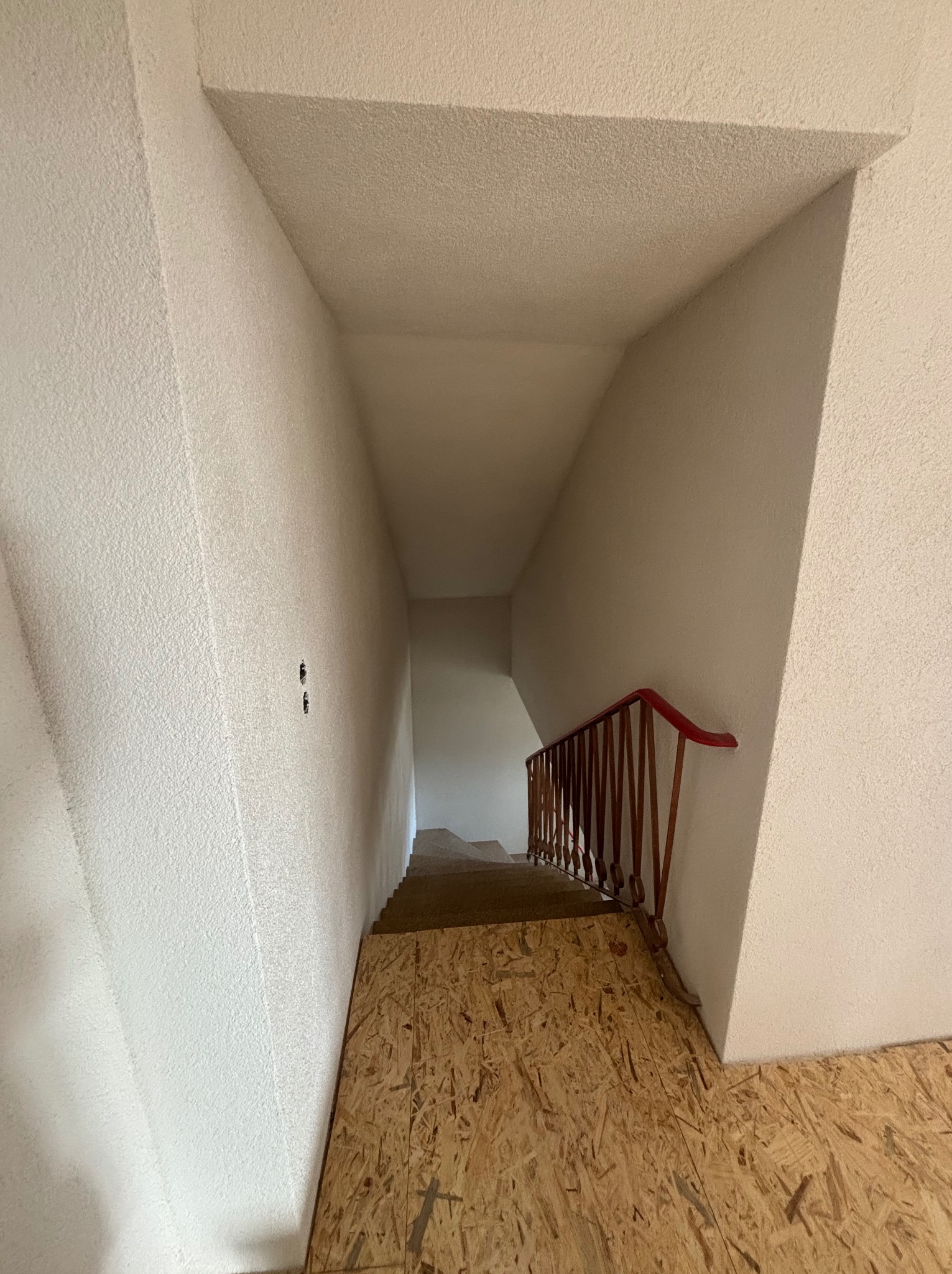 Schody w trakcie remontu: surowe ściany o fakturze baranka, tymczasowa podłoga z płyt OSB i drewniana balustrada z czerwoną poręczą. Widok z góry.