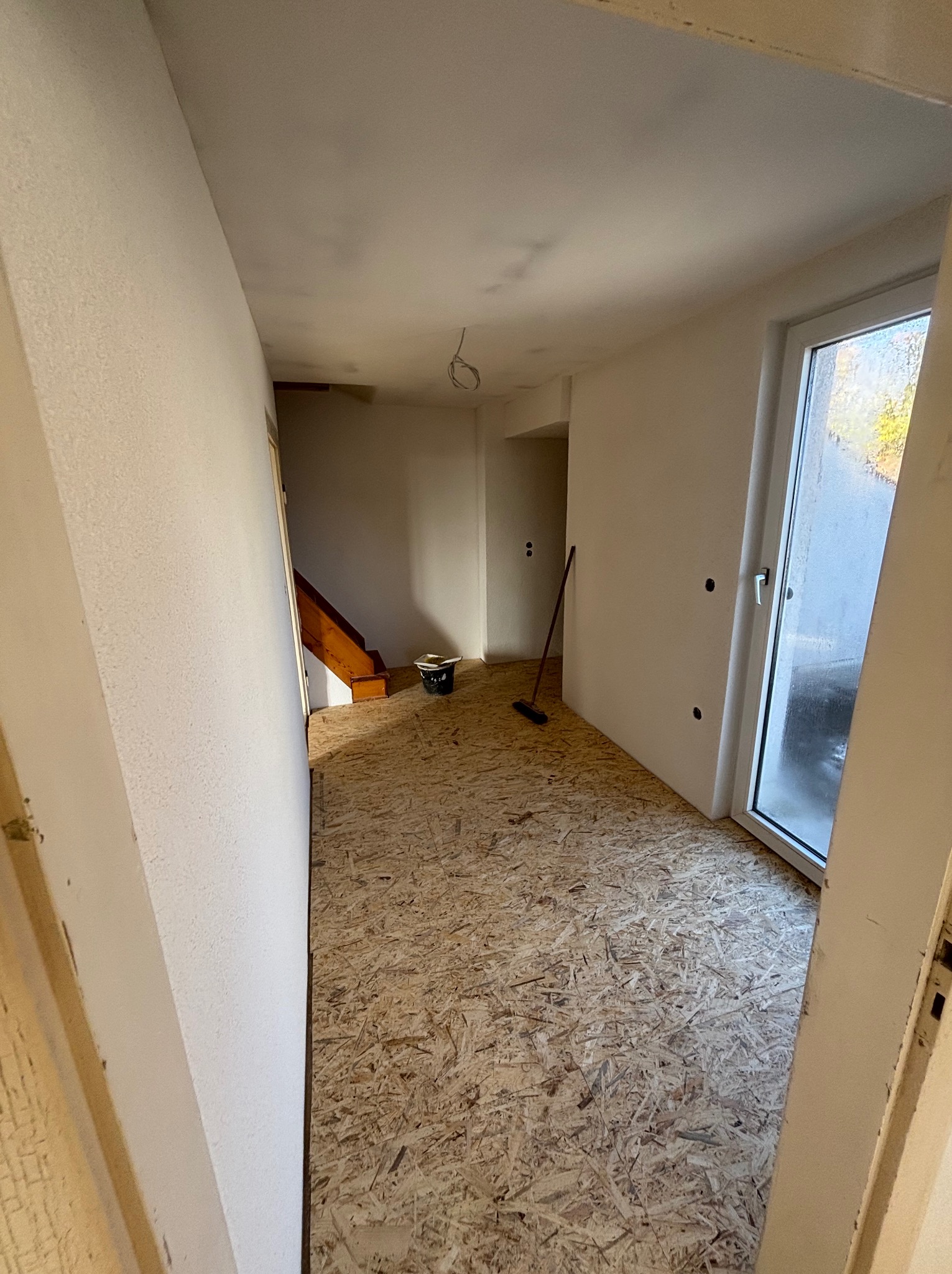 Wnętrze w trakcie remontu: surowe płyty OSB na podłodze, świeżo pomalowane ściany na biało, widoczne instalacje elektryczne i fragment schodów. Prace wykończeniowe w toku.