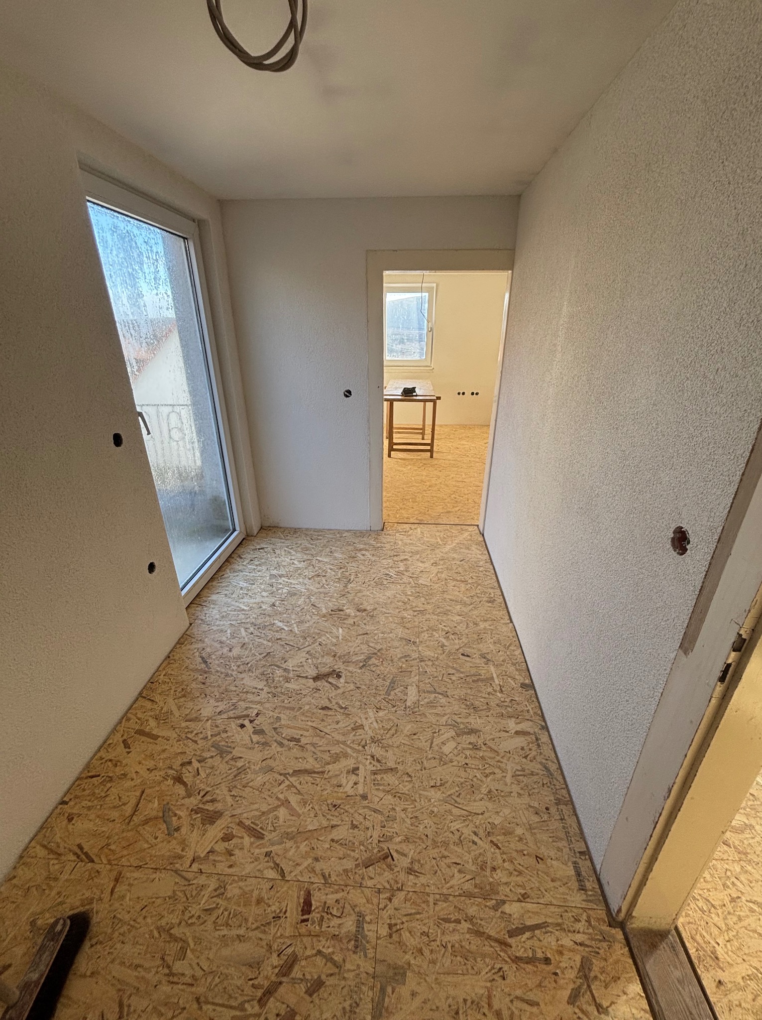 Wnętrze w trakcie prac wykończeniowych: surowe ściany z fakturą, podłoga z płyt OSB, widoczne kable i przygotowania do montażu drzwi. Perspektywa z korytarza.