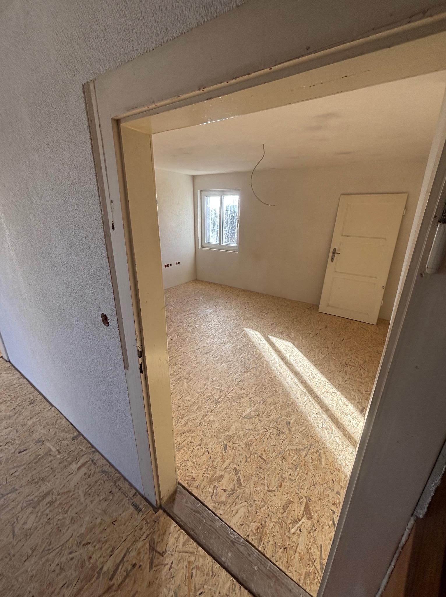 Wnętrze pokoju z podłogą wyłożoną płytami OSB, widok przez futrynę drzwiową. Ściany białe, okno z widokiem na zewnątrz. Przy ścianie białe drzwi.