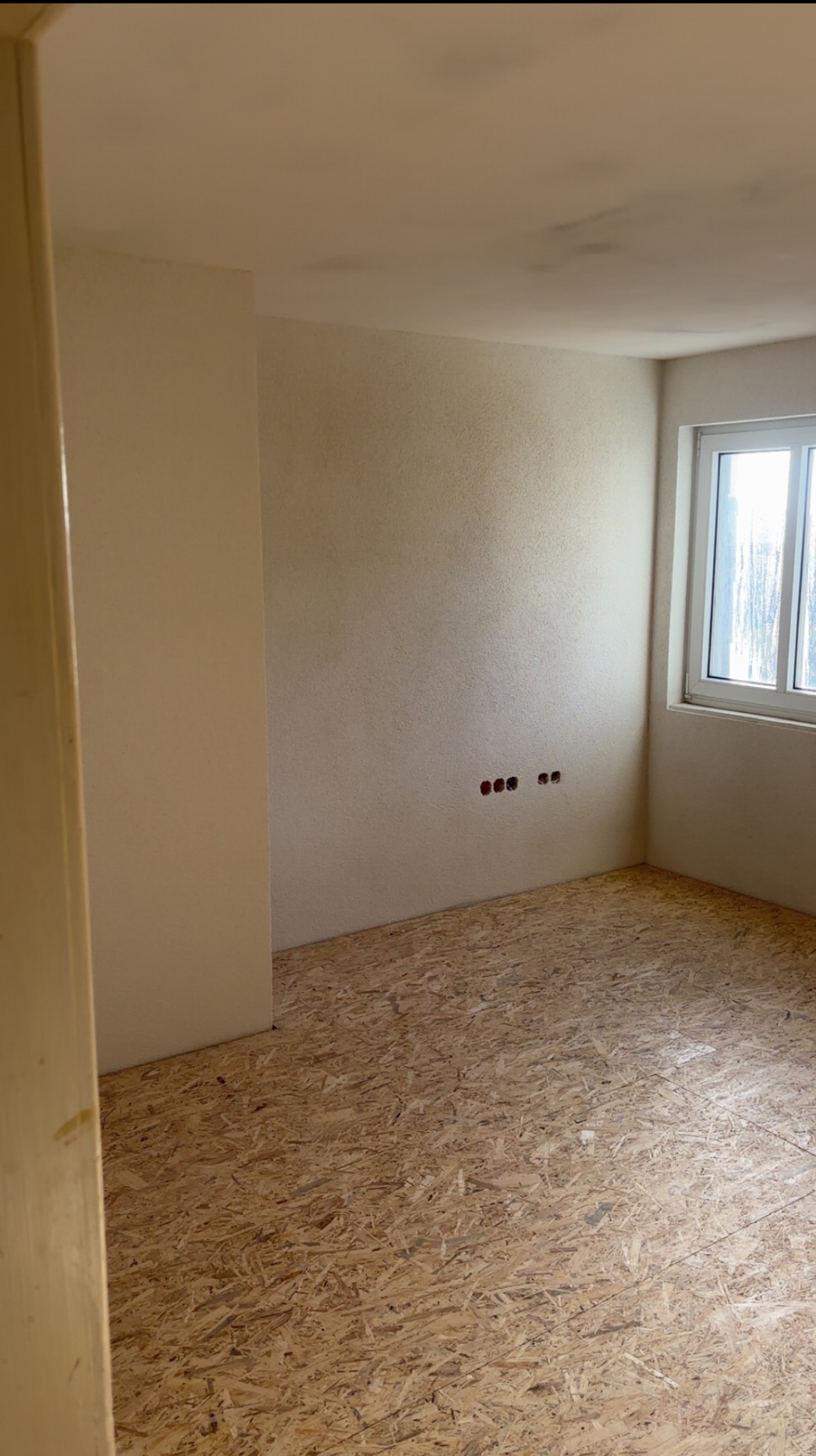 Surowa podłoga OSB w pokoju z oknem i gładzią na ścianach, przygotowana do dalszych prac wykończeniowych. Widoczne puszki elektryczne.