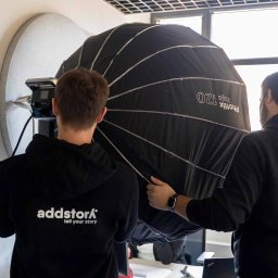 ADDSTORY SP. Z O.O. - Dwóch mężczyzn w bluzach z logo firmy ustawia lampę Phottix Raja 120 w studio z panelem akustycznym. Przygotowania do sesji zdjęciowej.