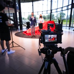 ADDSTORY SP. Z O.O. - Kamera na statywie rejestruje parę prezenterów na tle panoramicznego okna i czerwonej tkaniny. Operator kamery w tle po lewej.