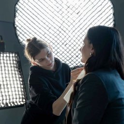 ADDSTORY SP. Z O.O. - Wizażystka poprawia makijaż klientce w studio, w tle lampy oświetleniowe. Kobieta w czarnej bluzie z kapturem delikatnie nakłada puder na twarz kobiety w marynarce.