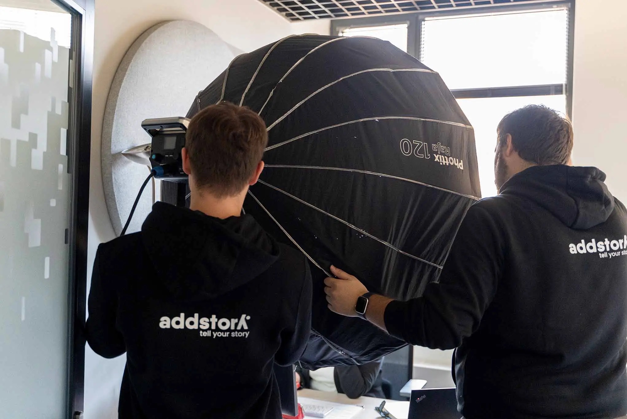 Dwóch mężczyzn w bluzach z logo firmy ustawia lampę Phottix Raja 120 w studio z panelem akustycznym. Przygotowania do sesji zdjęciowej.