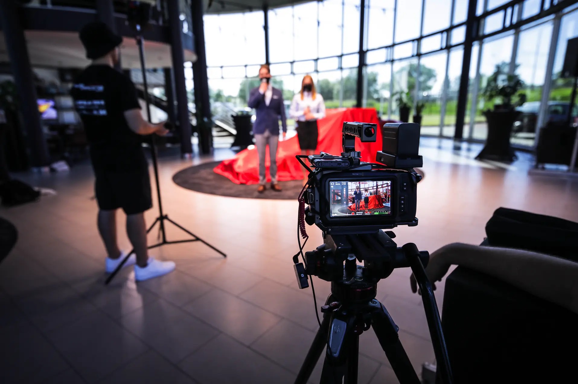 Kamera na statywie rejestruje parę prezenterów na tle panoramicznego okna i czerwonej tkaniny. Operator kamery w tle po lewej.