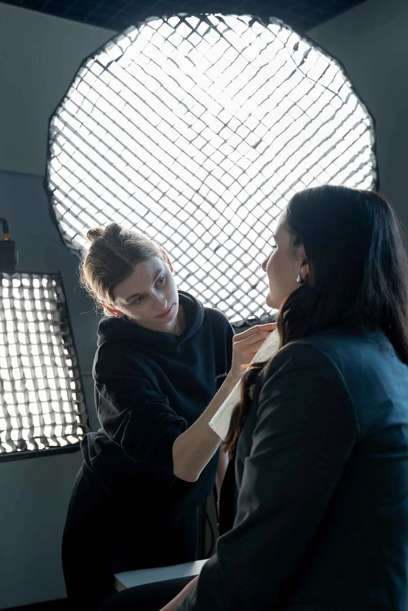 Wizażystka poprawia makijaż klientce w studio, w tle lampy oświetleniowe. Kobieta w czarnej bluzie z kapturem delikatnie nakłada puder na twarz kobiety w marynarce.