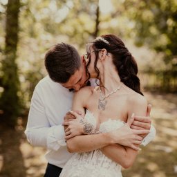 Mądre Kadry FotografSierakowice - Para młoda w objęciach na tle zieleni. Panna młoda w sukni z koronką, z tatuażami na ramionach i naszyjnikiem. Pan młody w białej koszuli i muszce całuje szyję kobiety.