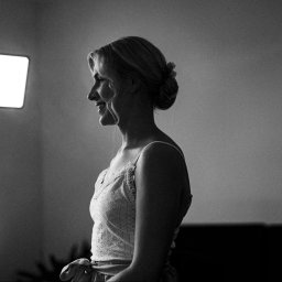 Mądre Kadry FotografSierakowice - Czarno-białe zdjęcie kobiety z profilu, podczas stylizacji włosów lakierem. Widoczne oświetlenie studyjne. Uczesanie upięte. Delikatna sukienka. Subtelny uśmiech.