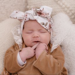 Mądre Kadry FotografSierakowice - Śpiąca noworodka w brązowym welurowym ubranku i opasce w kwiaty, leżąca na białym, puszystym kocyku. Palec przy ustach.