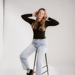 Mądre Kadry FotografSierakowice - Portret kobiety siedzącej na stołku w studio, ubrana w czarną bluzkę i jasne jeansy, z rękami uniesionymi za głową. Minimalistyczne tło, stonowana kolorystyka.