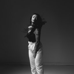 Mądre Kadry FotografSierakowice - Monochromatyczny portret kobiety z tatuażami, w czarnym topie i białych spodniach, z rozwianymi włosami na szarym tle. Dynamiczne ujęcie, podkreślające ruch i ekspresję.