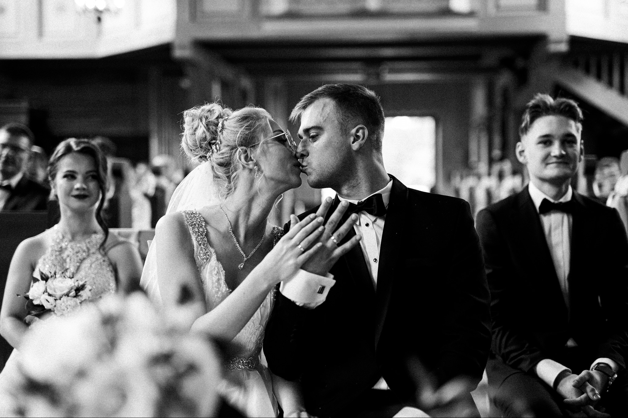 Czarno-białe zdjęcie z ceremonii ślubnej: panna młoda w okularach i pan młody w pocałunku, w tle goście. Panna młoda trzyma dłoń na ramieniu pana młodego.