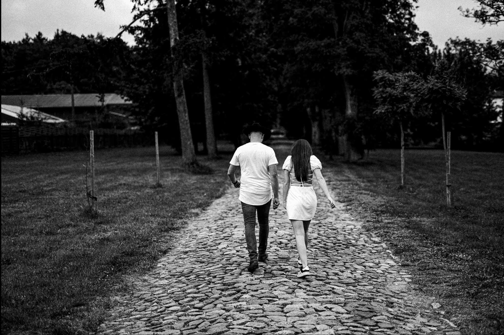 Czarno-białe zdjęcie pary idącej kamienistą ścieżką w parku, tyłem do obiektywu. Mężczyzna w kapeluszu i jeansach, kobieta w sukience z odkrytymi plecami. Drzewa i trawa po bokach.