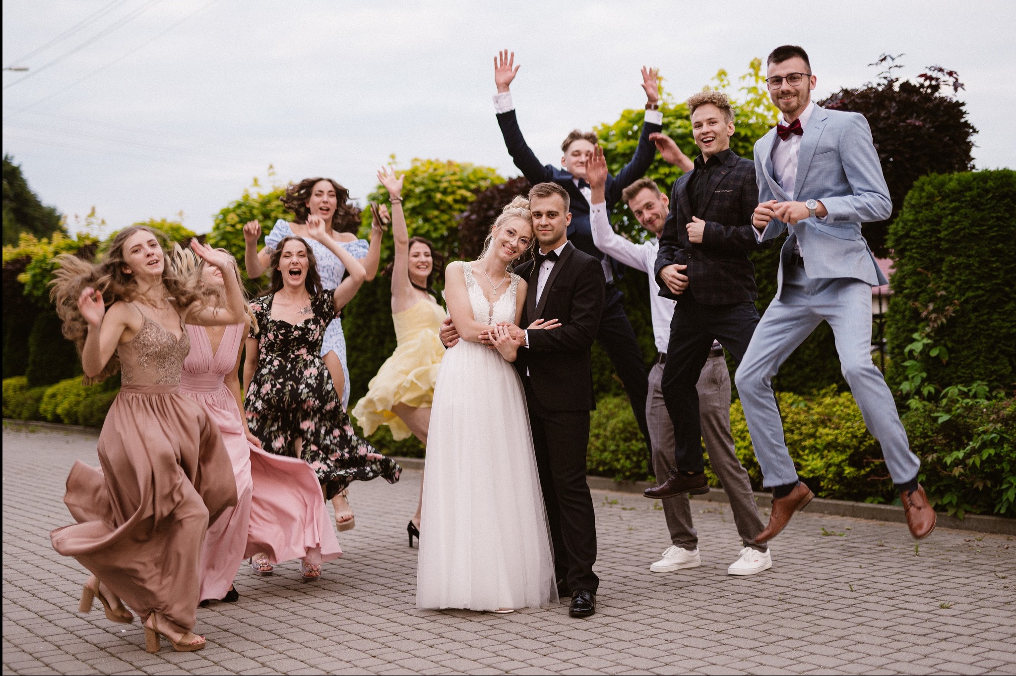 Radosna para młoda w otoczeniu skaczących gości weselnych na tle zieleni. Panna młoda w białej sukni, pan młody w czarnym garniturze. Dynamiczne ujęcie, energia i pozytywne emocje.