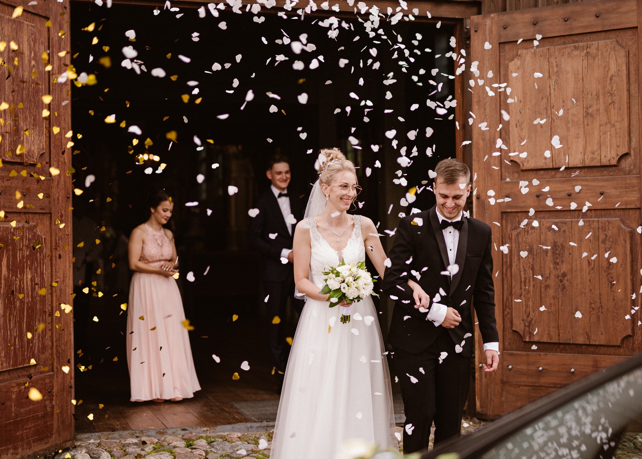 Uśmiechnięta para młoda wychodzi z drewnianych drzwi, obsypywana konfetti w kształcie serc. W tle goście weselni i fragment samochodu.