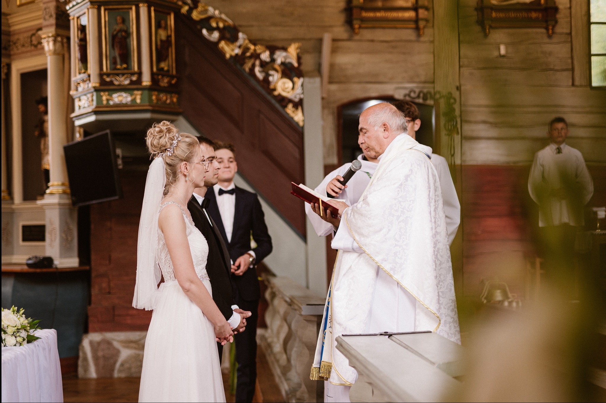 Uroczystość ślubna w kościele: Panna Młoda w białej sukni, obok Pan Młody w smokingu, ksiądz czyta z księgi, w tle drewniane ściany i schody.