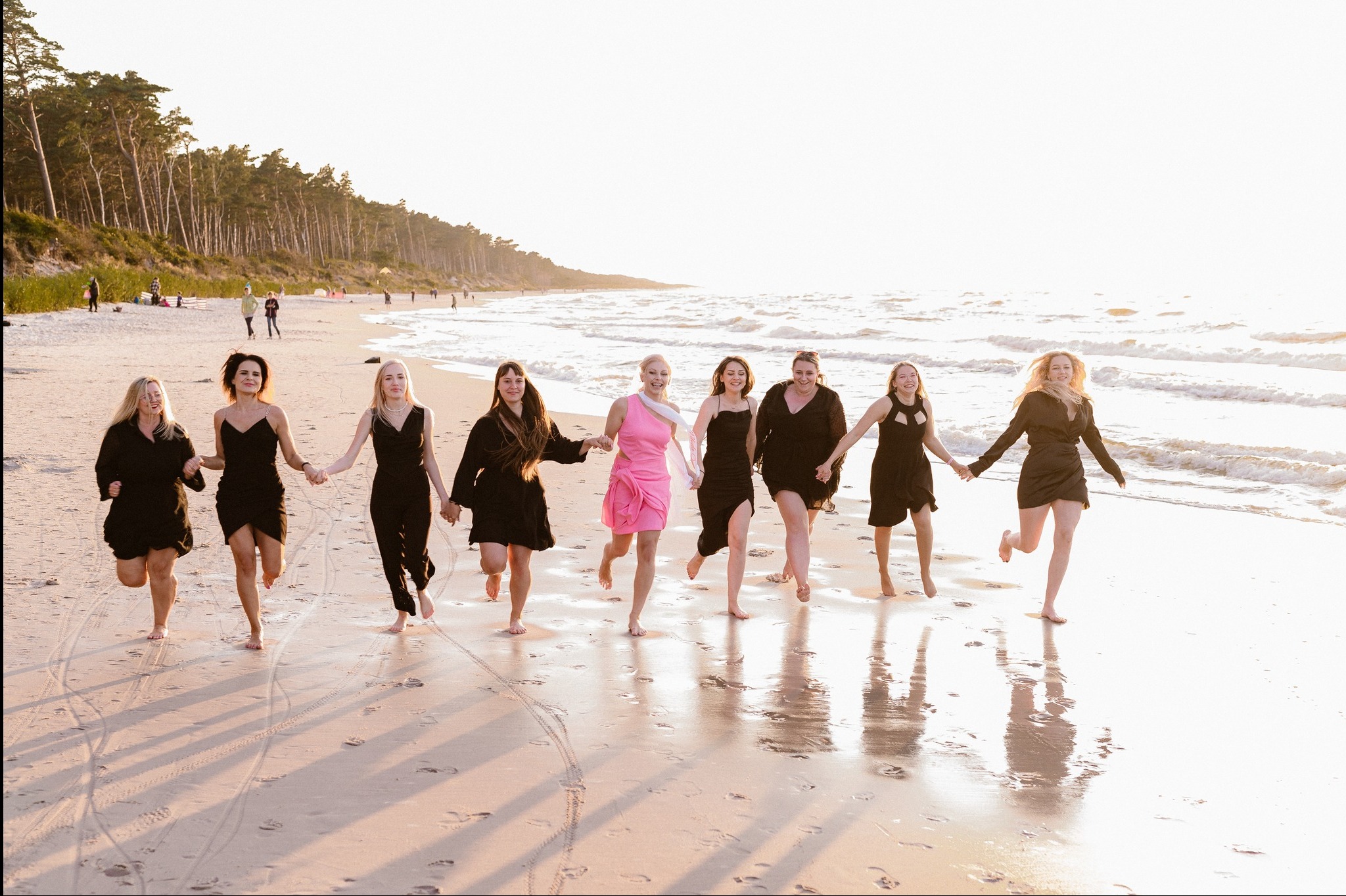 Grupa kobiet w sukienkach biegnie boso po plaży w słoneczny dzień. Jedna kobieta wyróżnia się różową sukienką. Widoczne ślady stóp na mokrym piasku i delikatne fale.