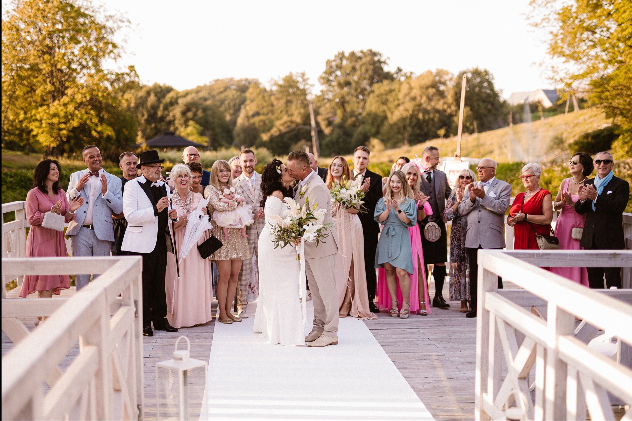 Para młoda całuje się na białym dywanie, otoczona gośćmi weselnymi na drewnianym pomoście, w tle drzewa i budynki. Uroczysta atmosfera.
