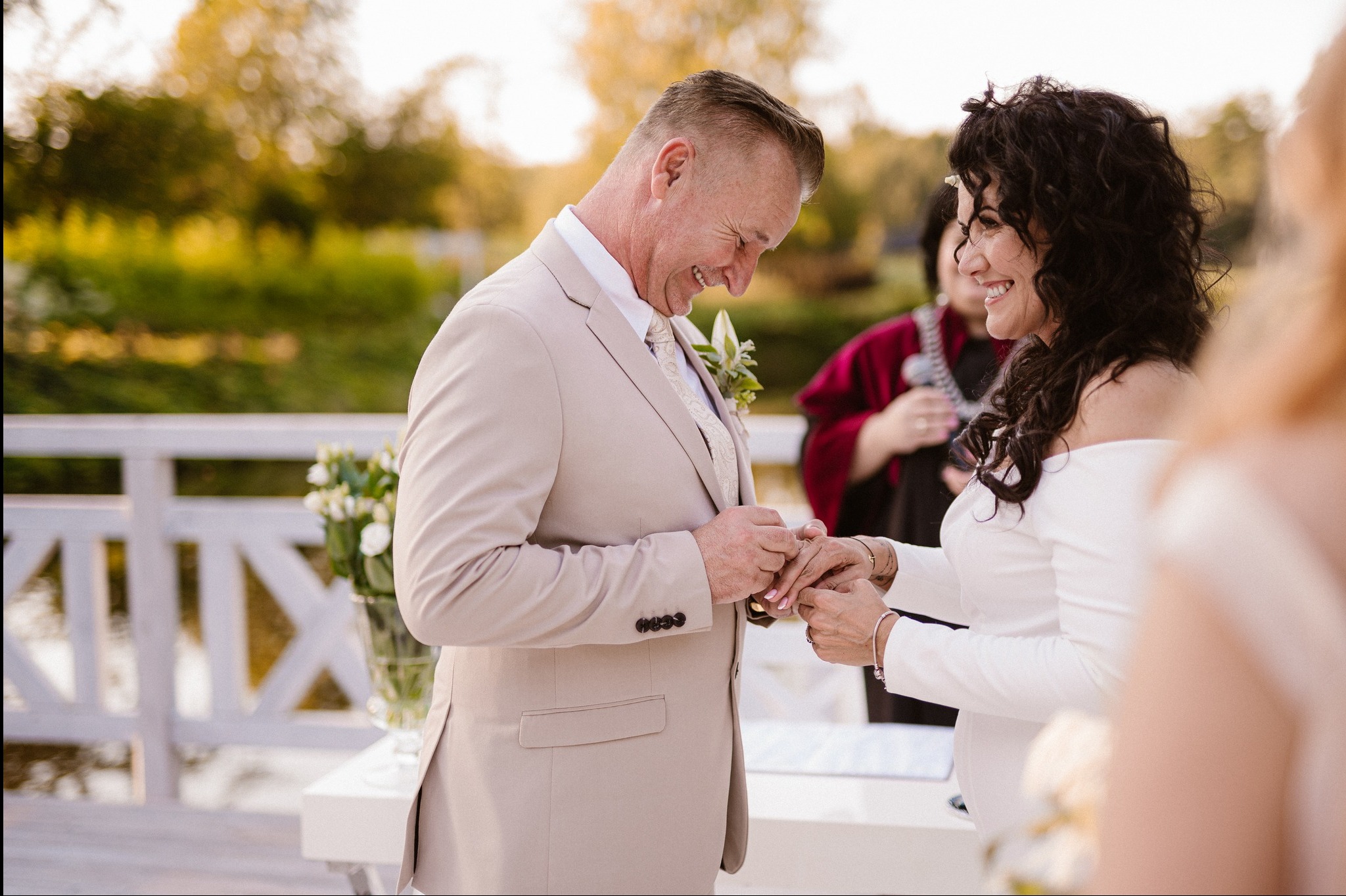 Uśmiechnięty pan młody w beżowym garniturze zakłada obrączkę pannie młodej w białej sukni, w tle kwiaty i goście podczas ceremonii ślubnej w plenerze.