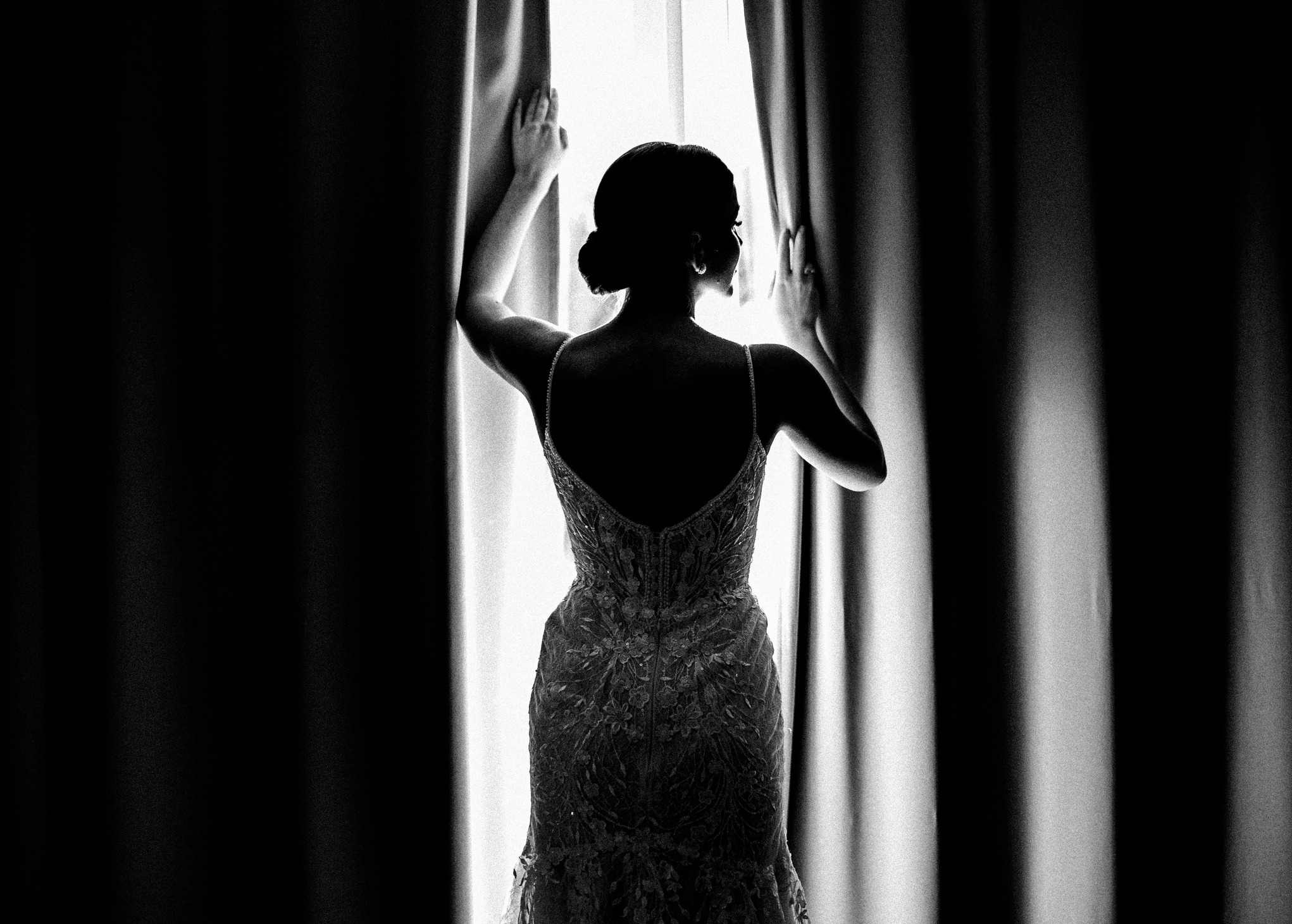 Elegancka sylwetka panny młodej w sukni z odkrytymi plecami, stojącej tyłem przy oknie z rozchylonymi zasłonami w czarno-białej tonacji.