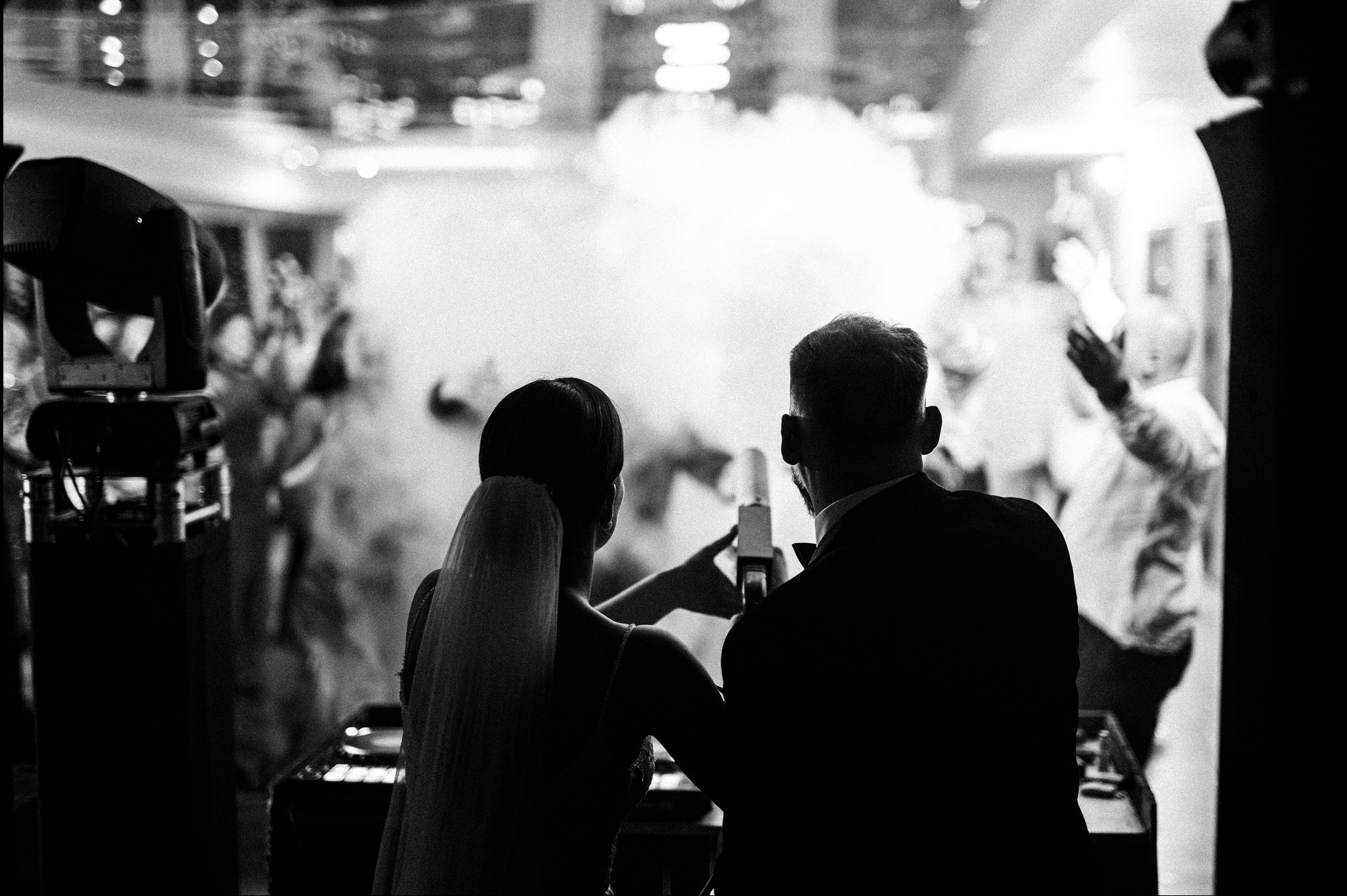 Czarno-białe ujęcie pary młodej od tyłu, z efektem dymu i oświetleniem scenicznym w tle, uchwycone podczas przyjęcia weselnego.