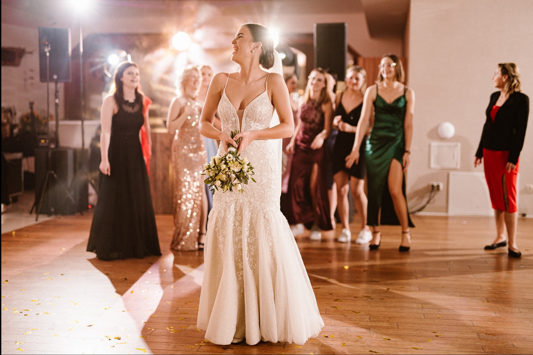 Uśmiechnięta panna młoda w koronkowej sukni, trzyma bukiet na parkiecie z konfetti, w tle druhny i oświetlenie sceniczne.