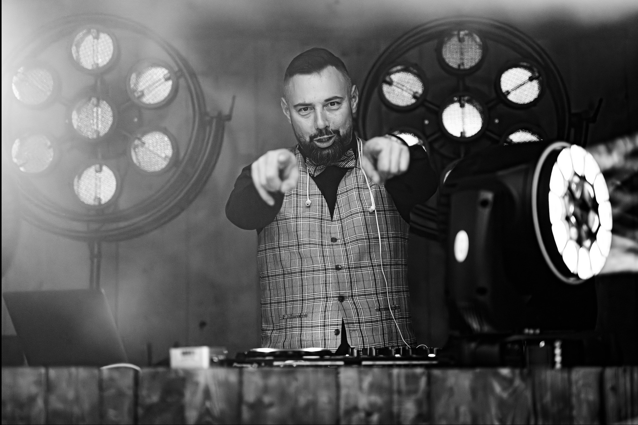 DJ w kraciastej kamizelce z muszką, wskazuje palcami w Sierakowicach, otoczony sprzętem i reflektorami. Czarno-białe ujęcie, dynamiczna poza.