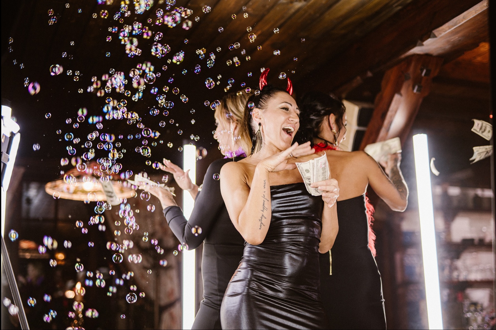 Uśmiechnięta kobieta w czarnej sukience z diabelskimi rogami trzyma plik banknotów na tle baniek mydlanych i dwóch innych kobiet.